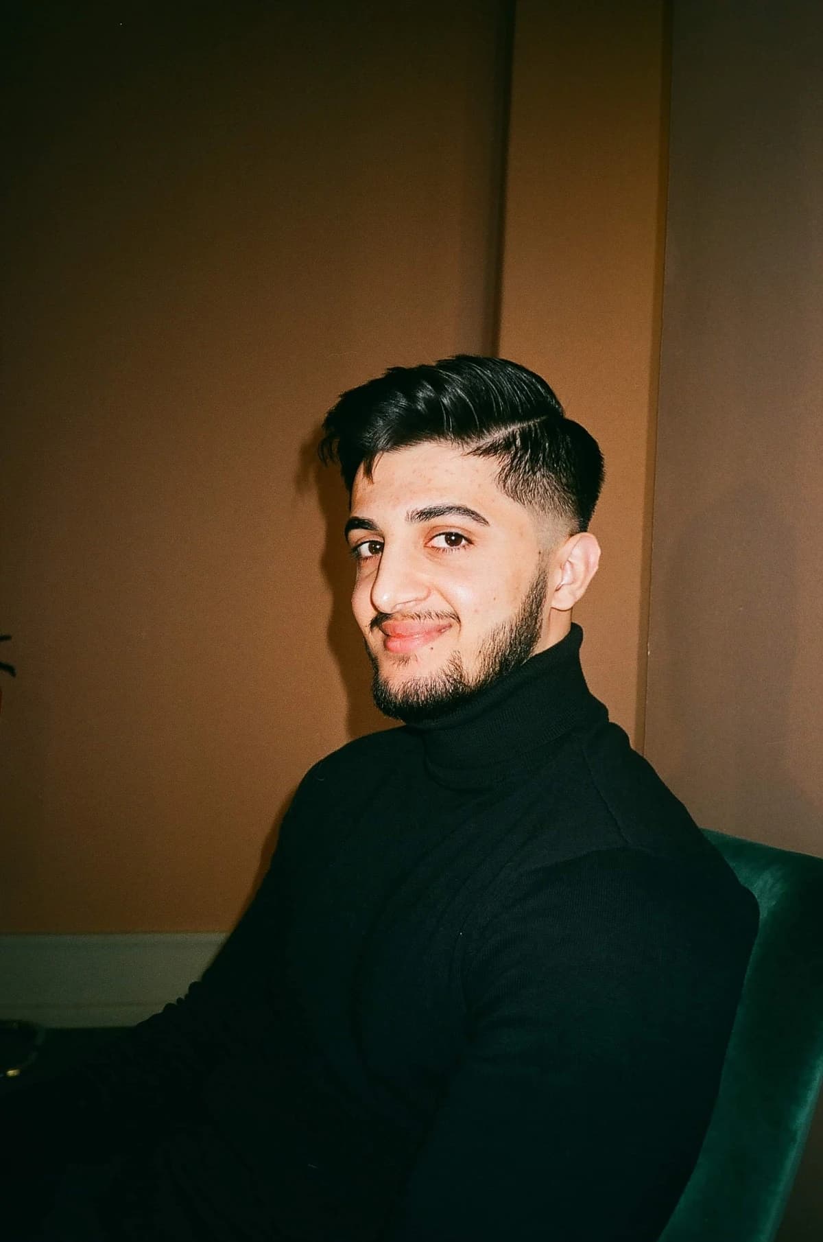 Daryan Kamalifar wearing a black turtleneck, seated beside a warm brown wall.